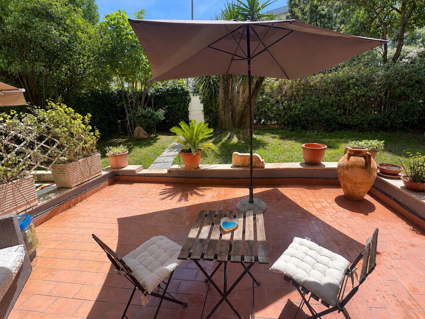 An outdoor patio with a table and chairs, surrounded by potted plants and a large umbrella.