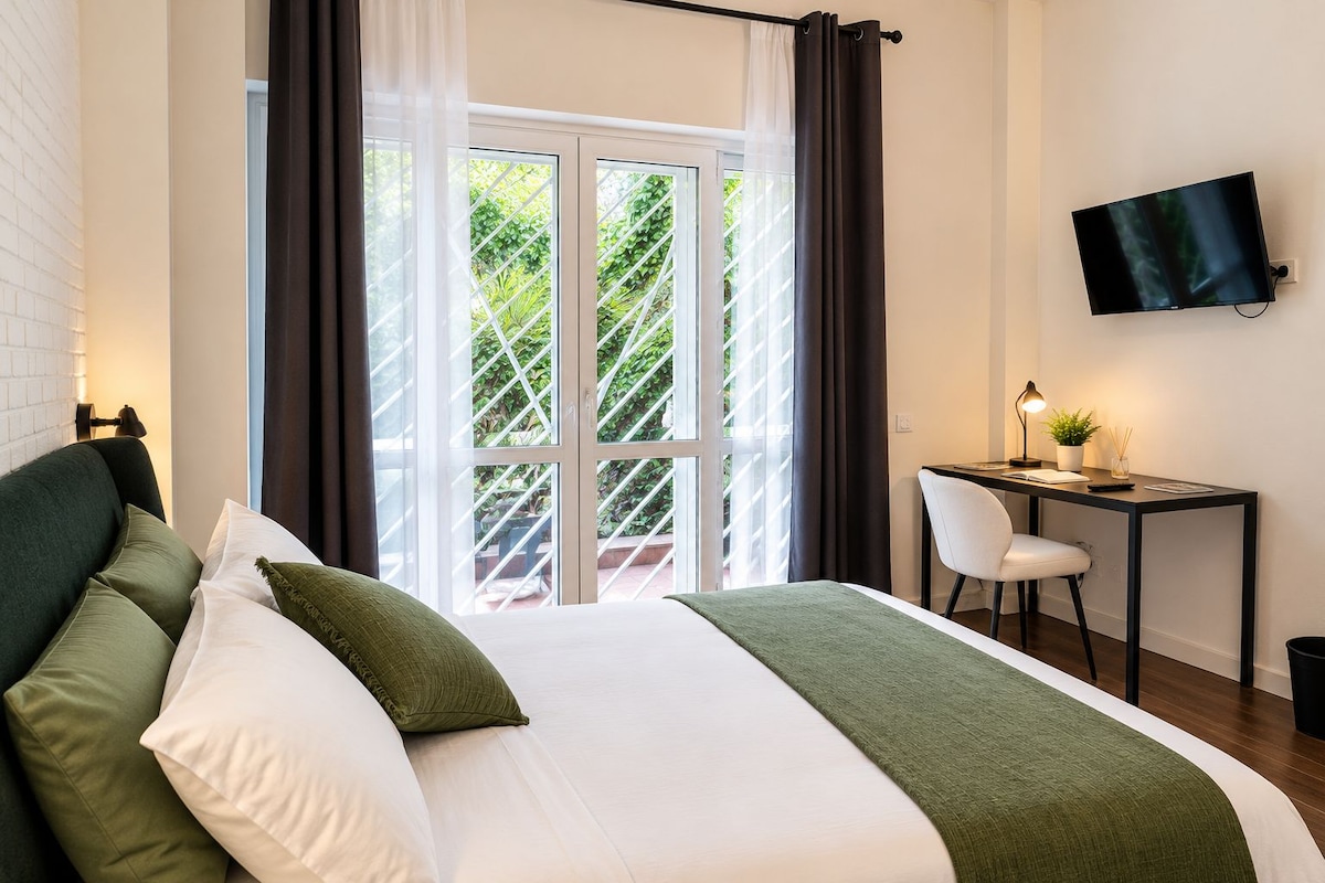 Quarto de hotel com cama, escrivaninha, cadeira, televisão e vista para o verde lá fora.