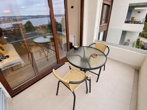 Ein Balkon mit Glasdoor, einem runden Glastisch, zwei Korbstühlen und Blick auf ein Gewässer und Gebäude.