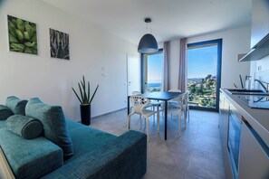 Un espacio habitable moderno con sofá, mesa de comedor, sillas, una planta en maceta y vistas a un paisaje costero a través de grandes ventanales.