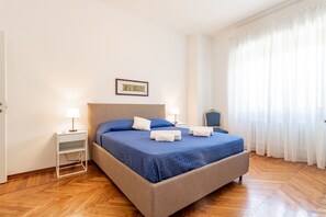 Ein Hotelzimmer mit Bett, Nachttisch, Lampe, Stuhl und einem Fenster mit Vorhängen.