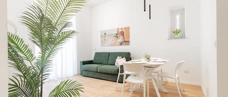 A modern living room with a green sofa, white dining table, and chairs, a large potted plant, and a painting on the wall.