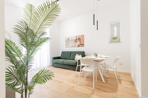 Een moderne woonkamer met een groene bank, een witte eettafel en stoelen, een grote potplant en een schilderij aan de muur.
