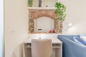 A room with a white desk, a chair, a blue couch, a stone wall with a brick arch, and a small plant. - ACQUAMARINA - Centro (Sanremo)