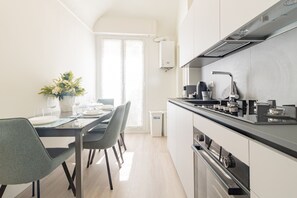 A modern kitchen with a dining area, featuring a stainless steel range hood, a coffee machine, and a trash bin. - ACQUAMARINA - Centro (Sanremo)