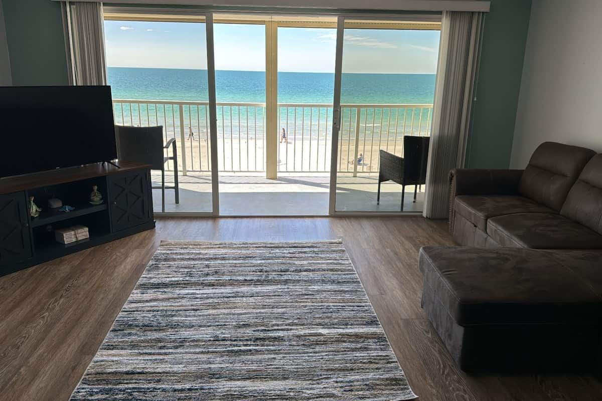 Una habitación con un amplio ventanal corredero que da a vistas de la playa, un televisor de pantalla plana, un sofá de cuero marrón y suelo de madera.