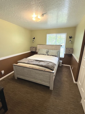 A bedroom with a wooden bed, a nightstand, a wall-mounted lamp, and a window with blinds. - Wasatch Chalet - 3BR Sandy Mountain Retreat (Sandy)