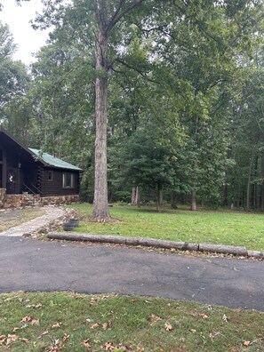 A log cabin with a stone foundation, surrounded by a grassy area and trees.
