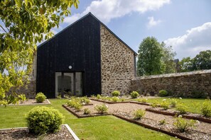 Ein Gebäude mit dunkler Fassade und Steinwand, umgeben von einem gepflegten Garten mit verschiedenen Pflanzen und blauem Himmel.
