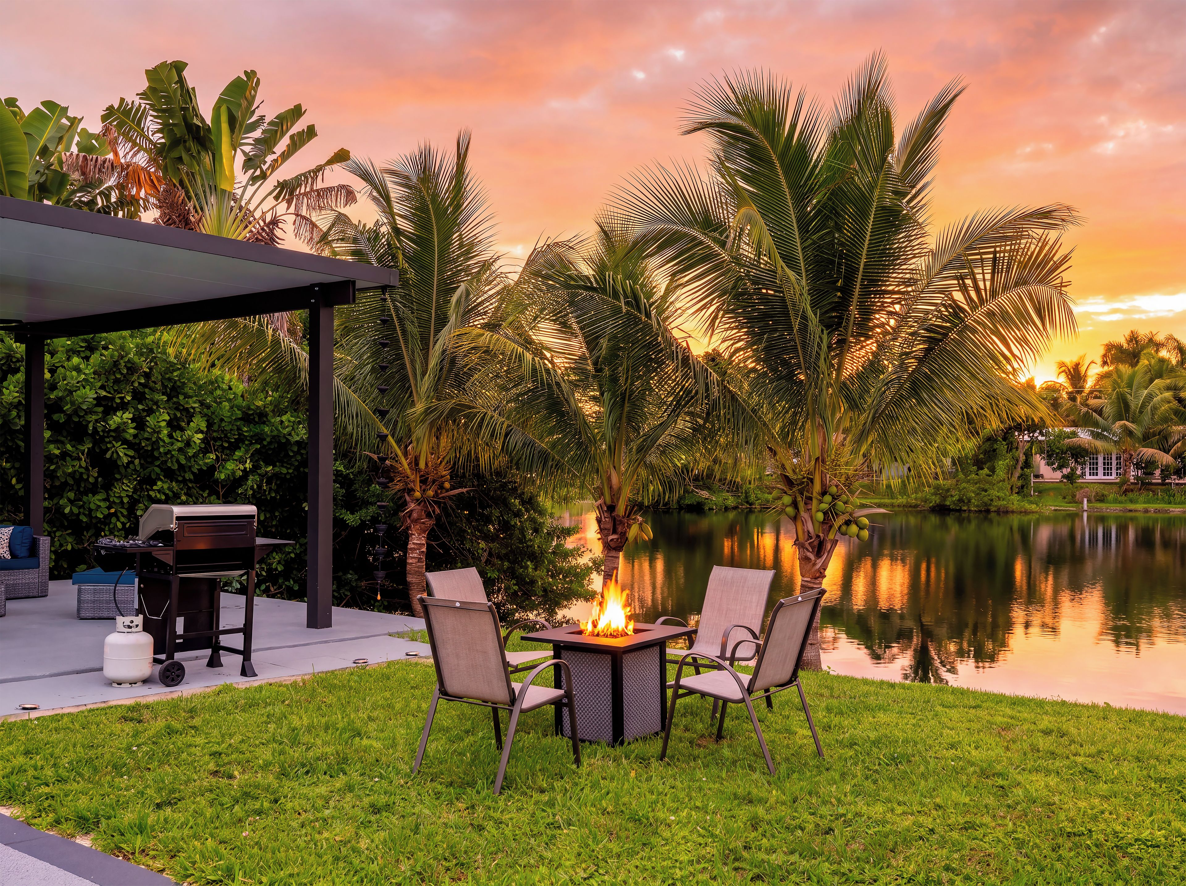 Eine Terrasse mit Grill, Stühlen und Feuerstelle am Ufer eines ruhigen Sees, Palmen und Sonnenuntergang.