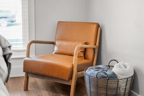 Ett hörnrum med en brun läderfåtölj, en korg av rotting med en kudde i och ett fönster med persienner.