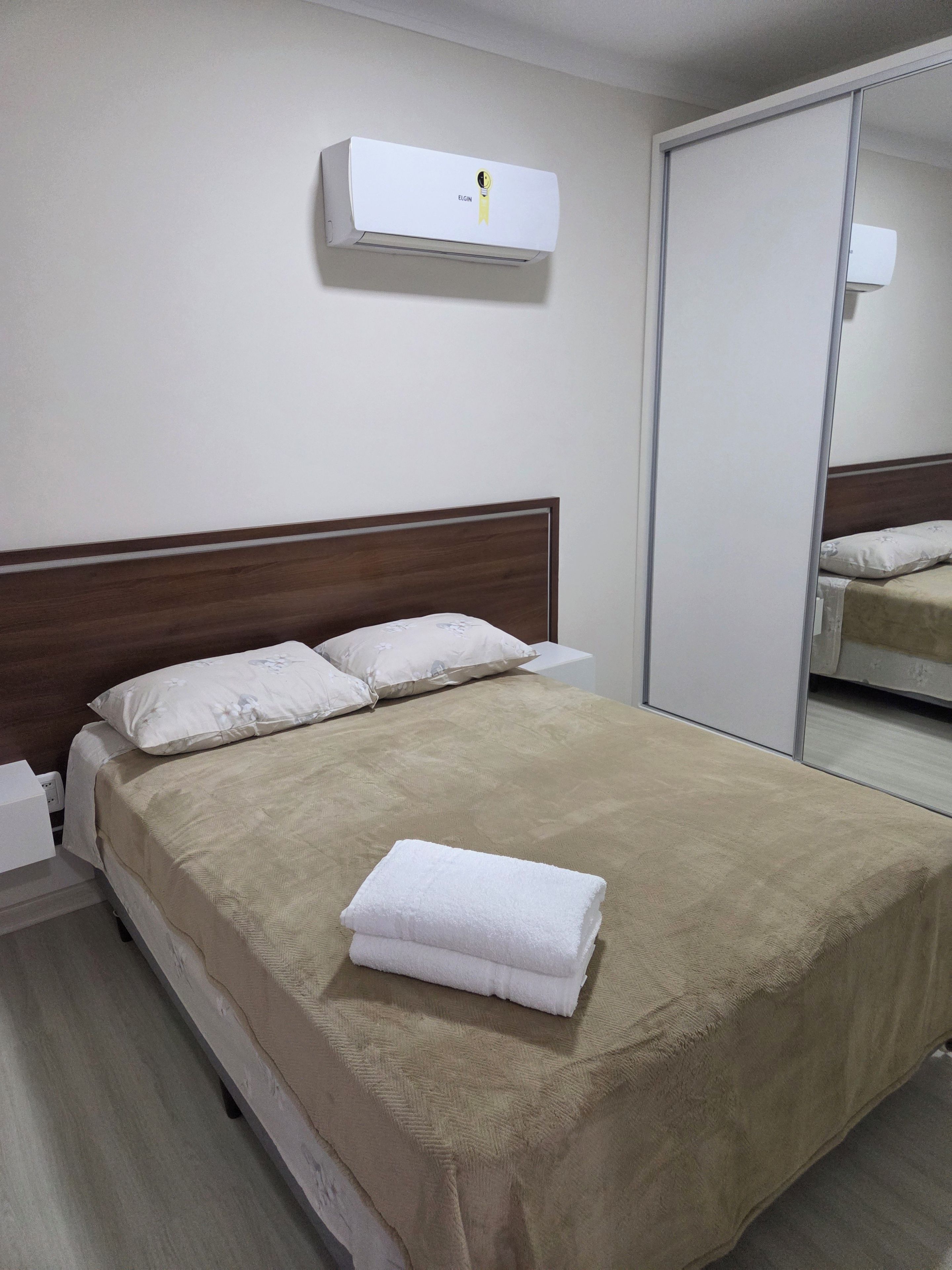 Ein ordentlich bezogenes Bett mit einer beigen Decke, weißen Kissen und einem gefalteten weißen Handtuch.