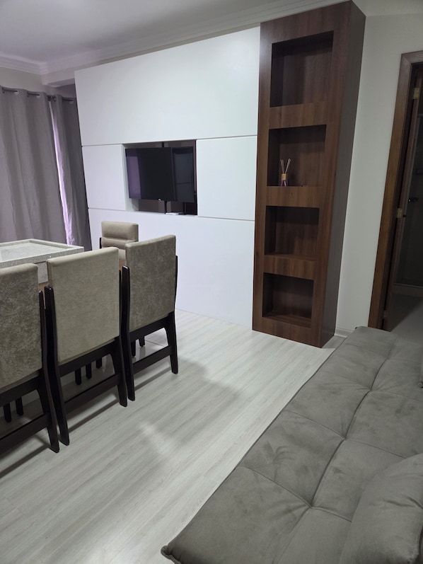 A modern living room with a sofa, a dining table with chairs, a TV mounted on the wall, and a wooden cabinet.