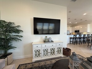 A living room with a flat-screen TV, a white cabinet, a large plant, and a kitchen area in the background. - Las Olas 304 -  Exclusive 3br Condo South Padre Island - Steps from the Sand (South Padre Island)