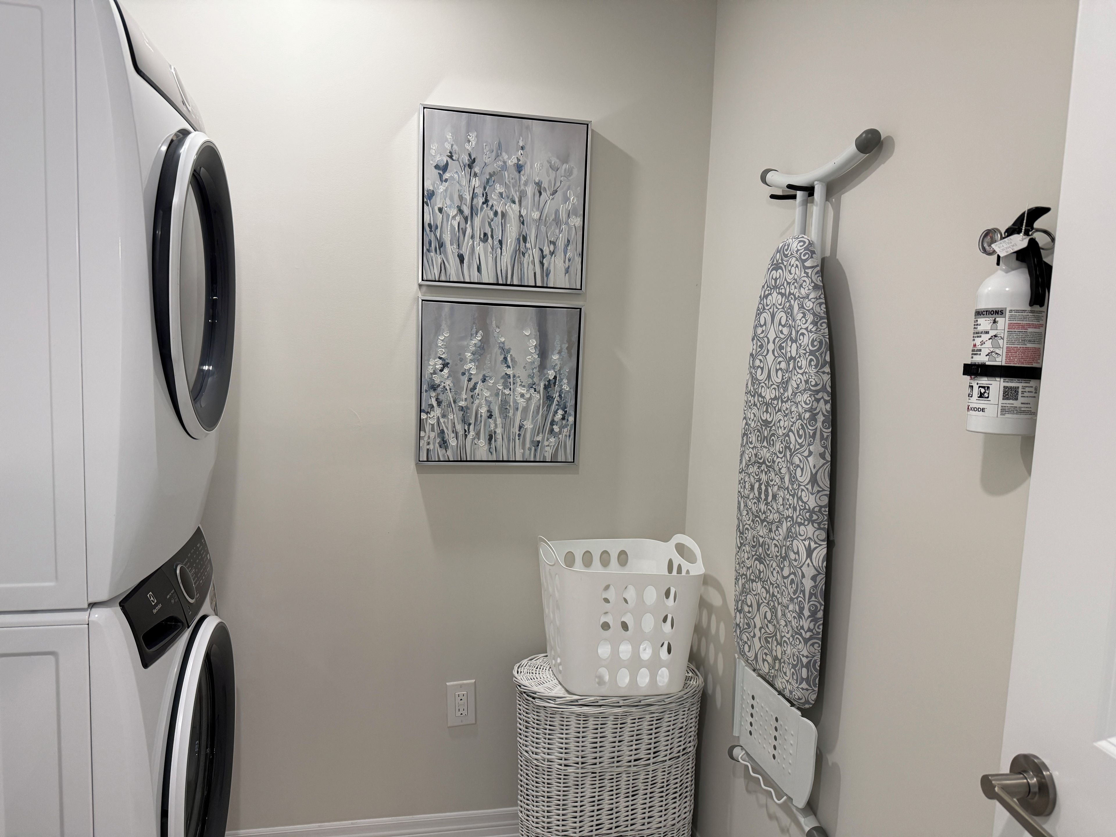 A laundry room with a washing machine, two framed artworks, a drying rack, and a basket.
