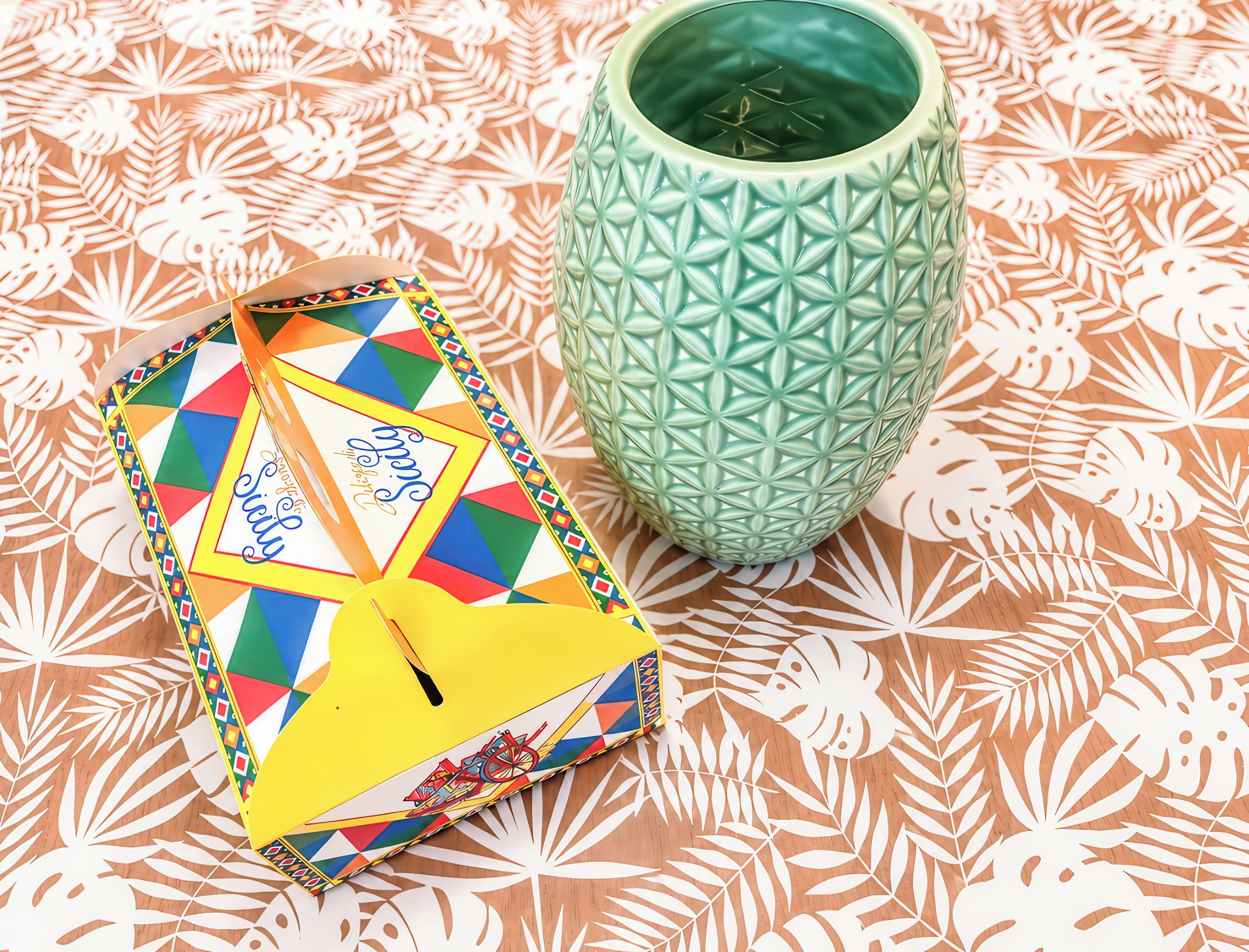 A colorful notebook with a geometric pattern and a green textured vase on a patterned tablecloth.
