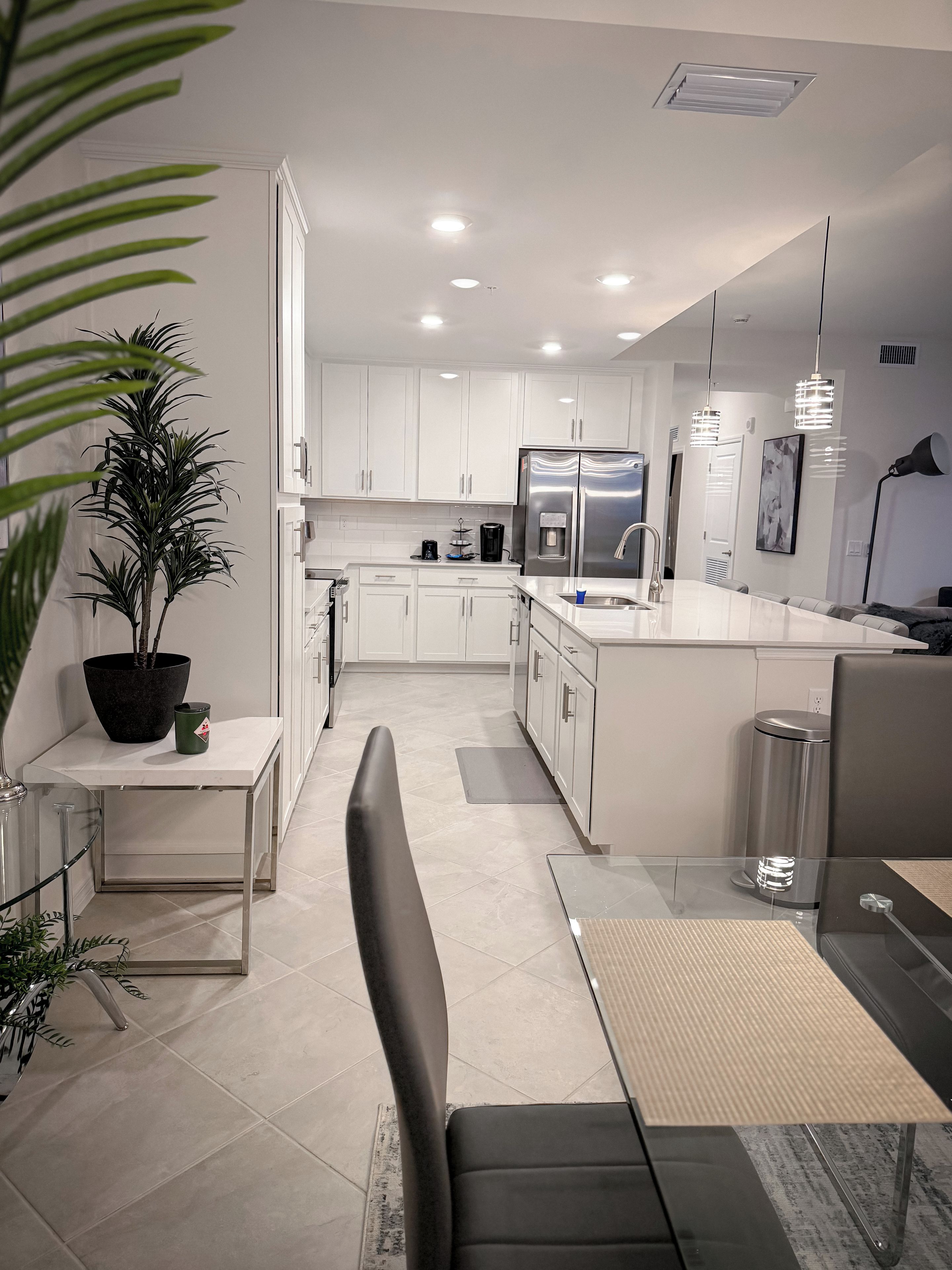Una cocina moderna con armarios blancos, un refrigerador de acero inoxidable y un comedor con mesa de vidrio y sillas negras.