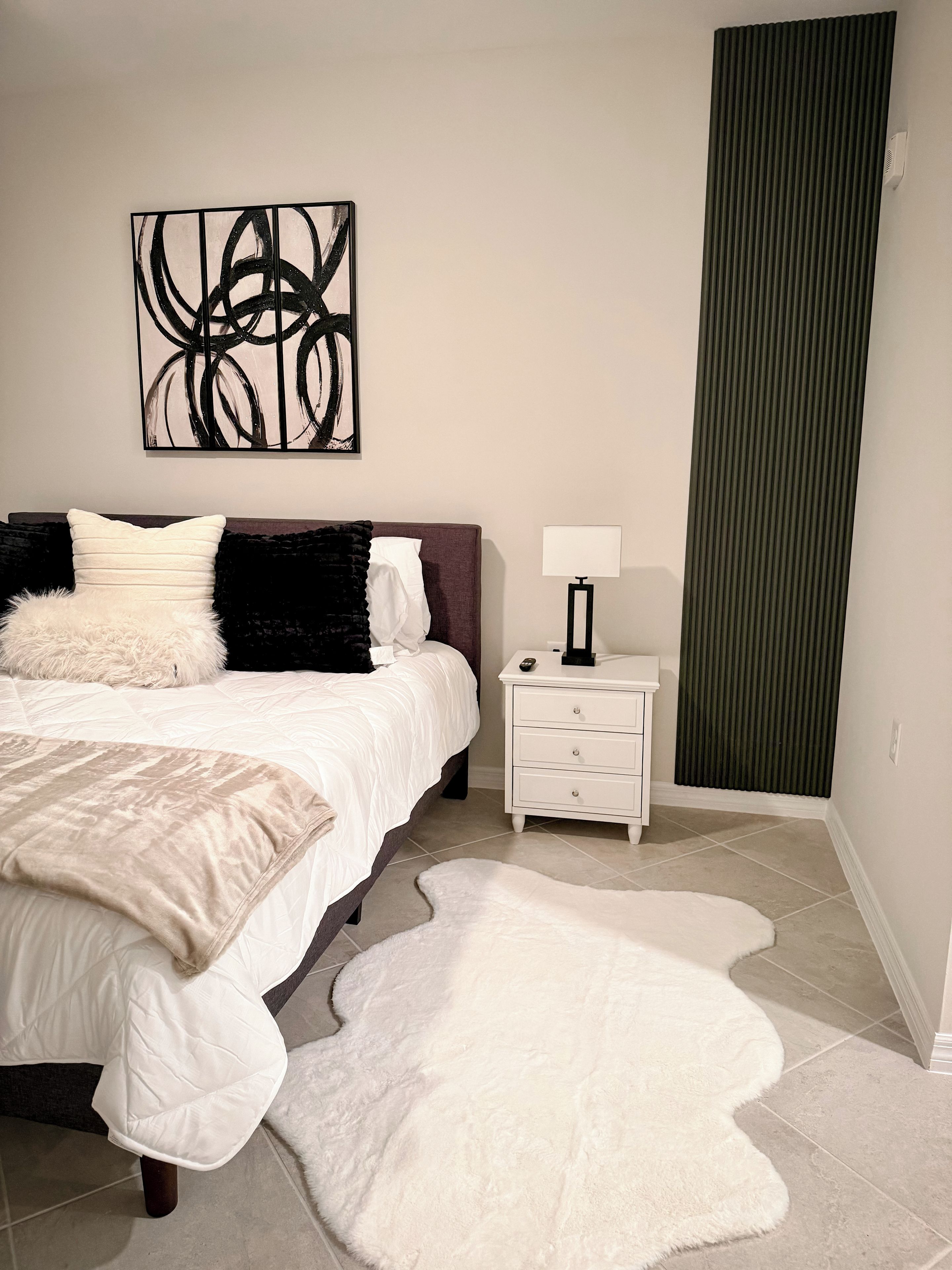 A bedroom with a bed, a nightstand, a lamp, a large abstract painting, and a green vertical panel.