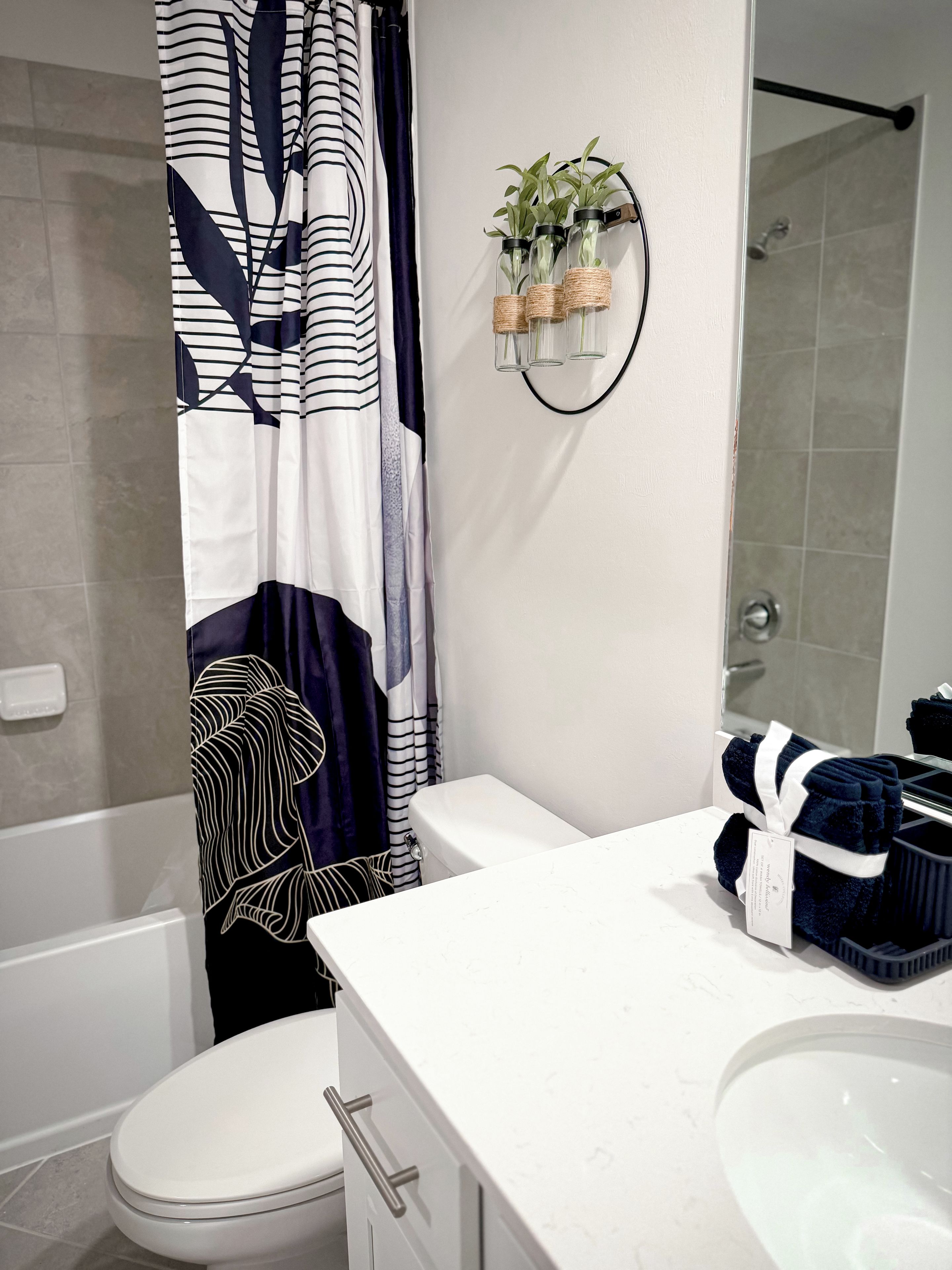 Une salle de bain avec des toilettes blanches, un lavabo blanc, un espace douche et un porte-serviettes agrémenté d’une plante.