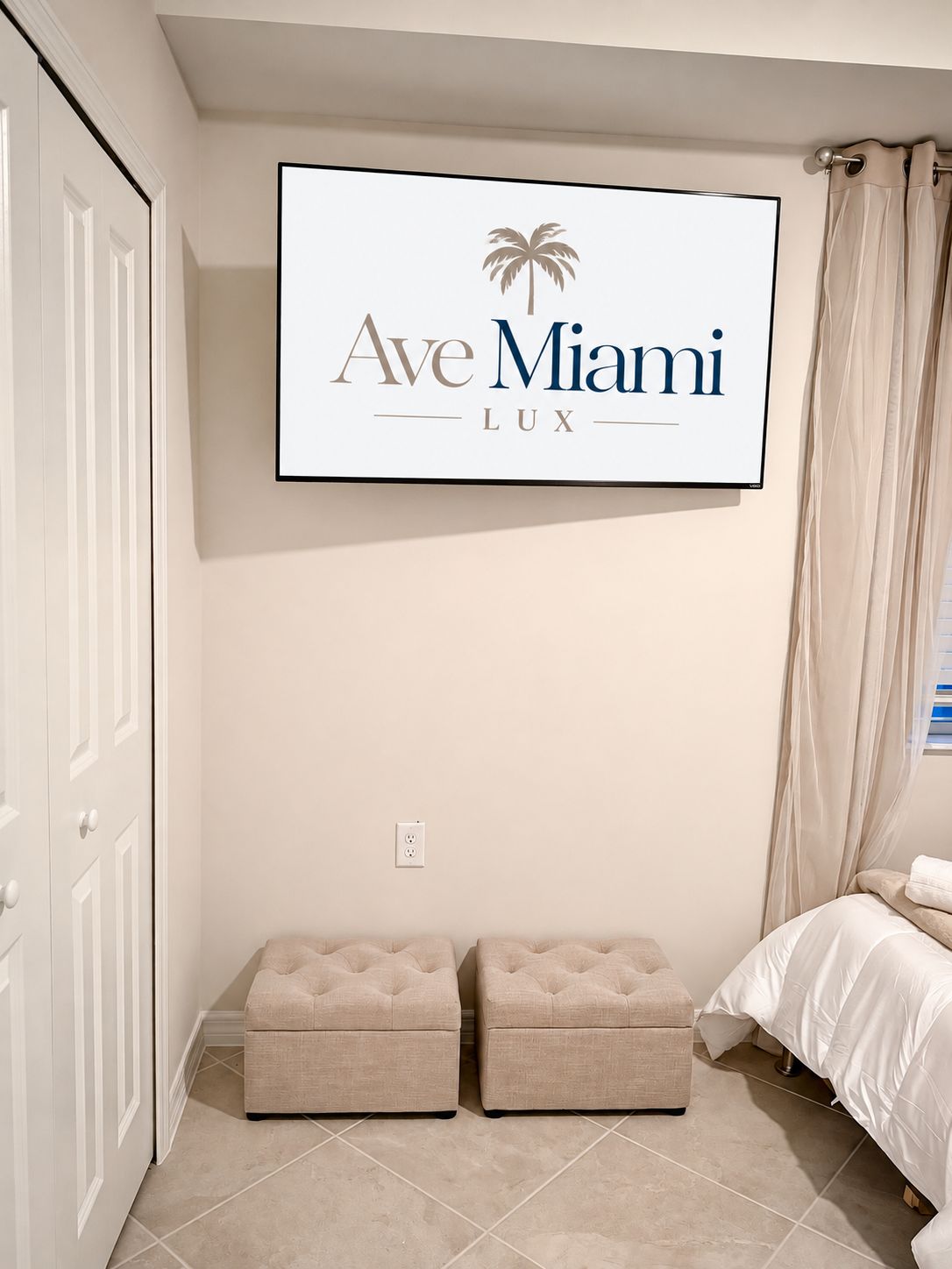 Una habitación de hotel con una cama, un televisor montado en la pared y dos otomanas.
