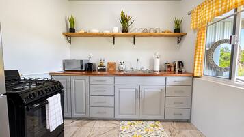 Una cocina con estufa, microondas y diversos electrodomésticos en la encimera. Hay armarios con cajones y una ventana con cortinas.