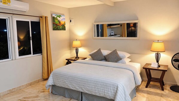Habitación de hotel con cama, dos mesitas de noche, ventilador de pie, ventana con cortinas y espejo montado en la pared.