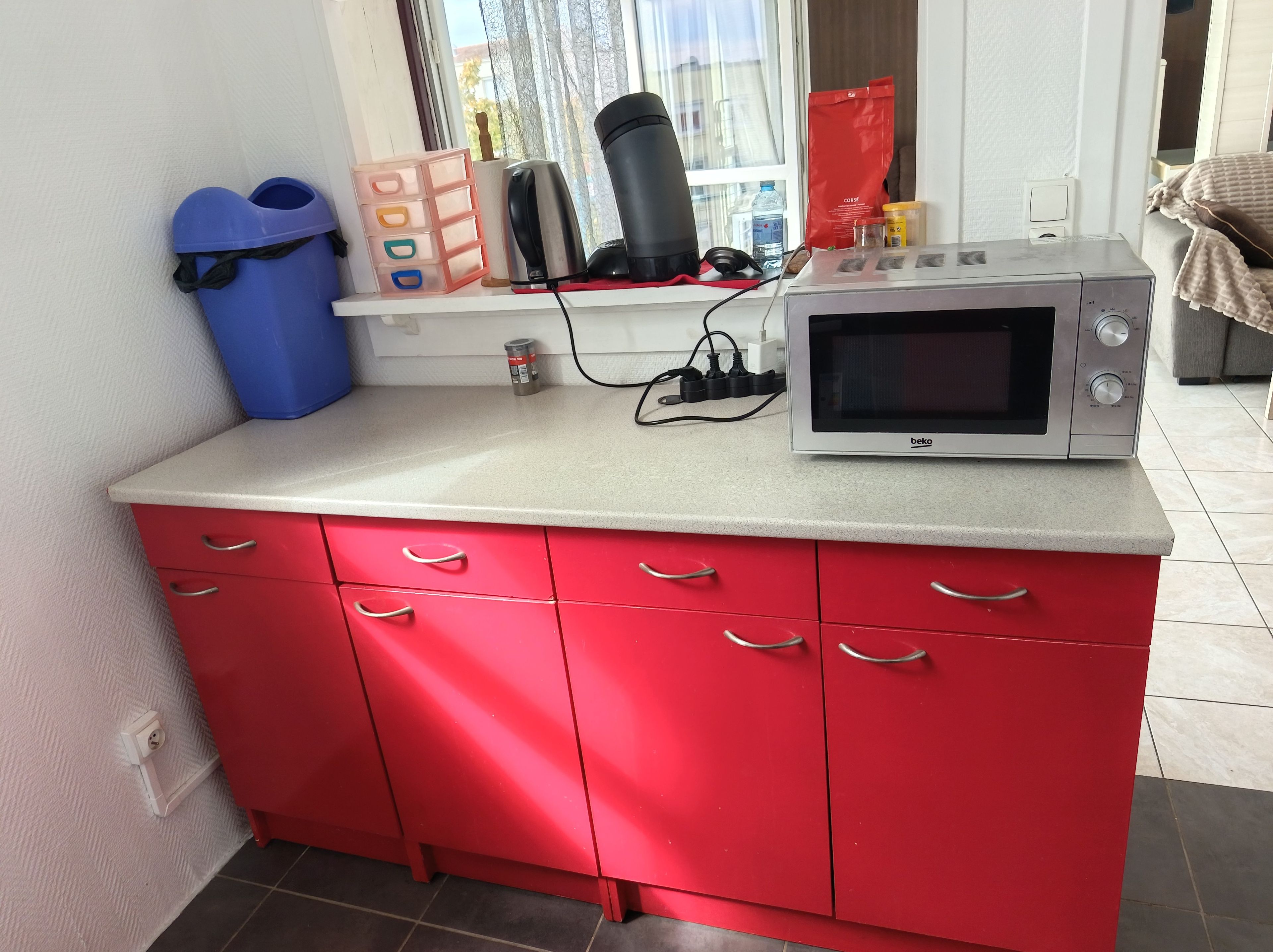 Une petite cuisine équipée d’un four à micro-ondes, d’un meuble rouge, d’une fenêtre et d’une poubelle bleue.