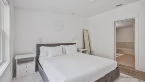 A bedroom with a bed, bedside tables, a nightstand, a mirror, and a door leading to a bathroom. - Beautifully updated East Hampton retreat with a heated pool, sauna, and... (East Hampton)