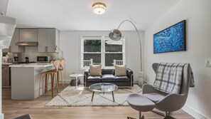 A modern kitchen with white cabinets, a dining area with a round glass table, and a living room with a sofa and armchair. - Beautifully updated East Hampton retreat with a heated pool, sauna, and... (East Hampton)