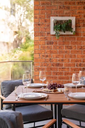 A dining table set with plates, glasses, and a bowl of grapes, against a brick wall with a hanging planter. - Manly 2BR | 5 Min Walk to Beach + Parking (Manly)