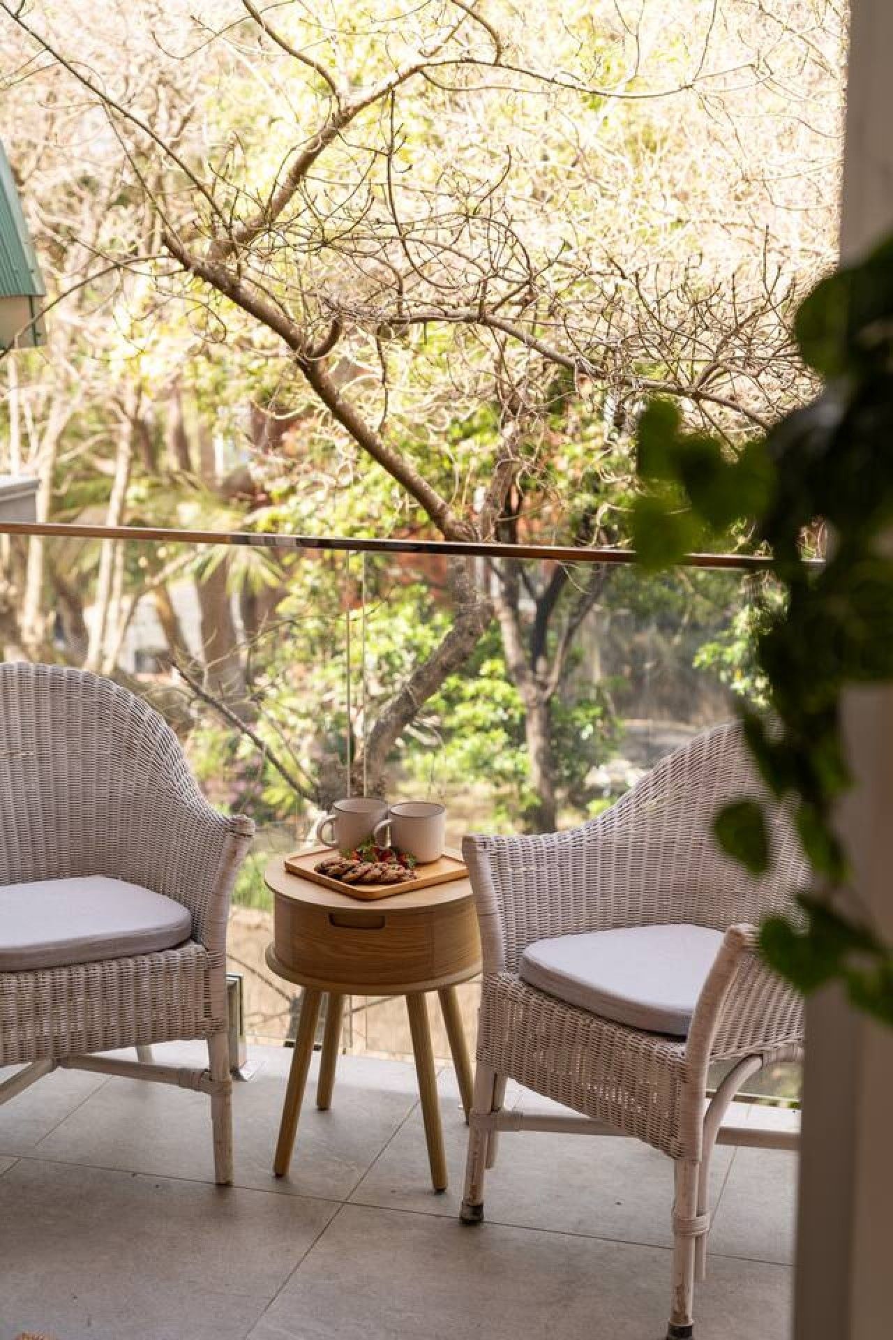 A balcony with wicker furniture, a small table with a tray of food, and a view of trees and greenery.