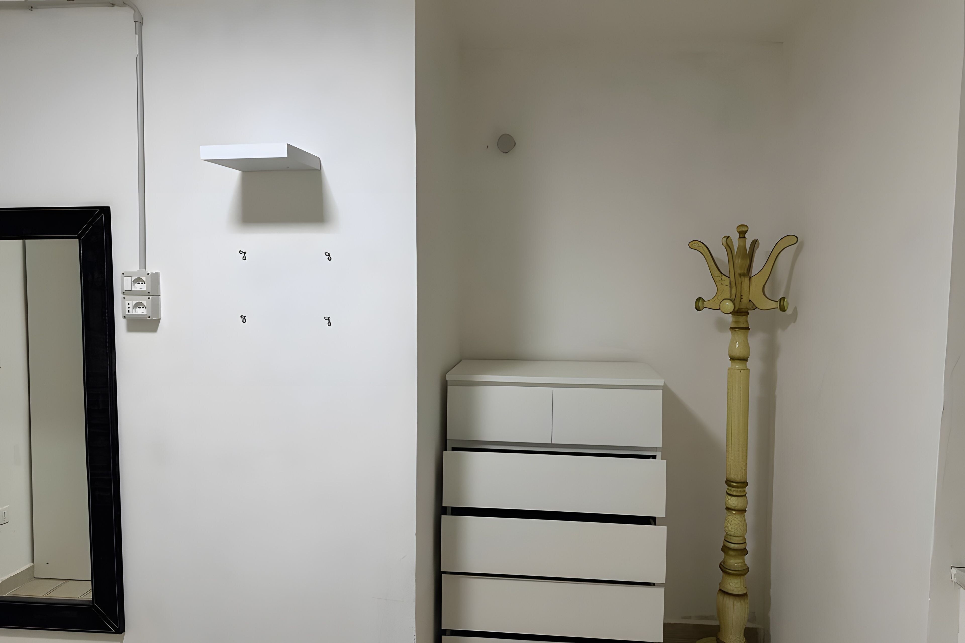 Ein Zimmer mit weißer Wand, einem Spiegel, einer Wandleuchte, einer weißen Kommode mit Schubladen und einer dekorativen Holzstange.