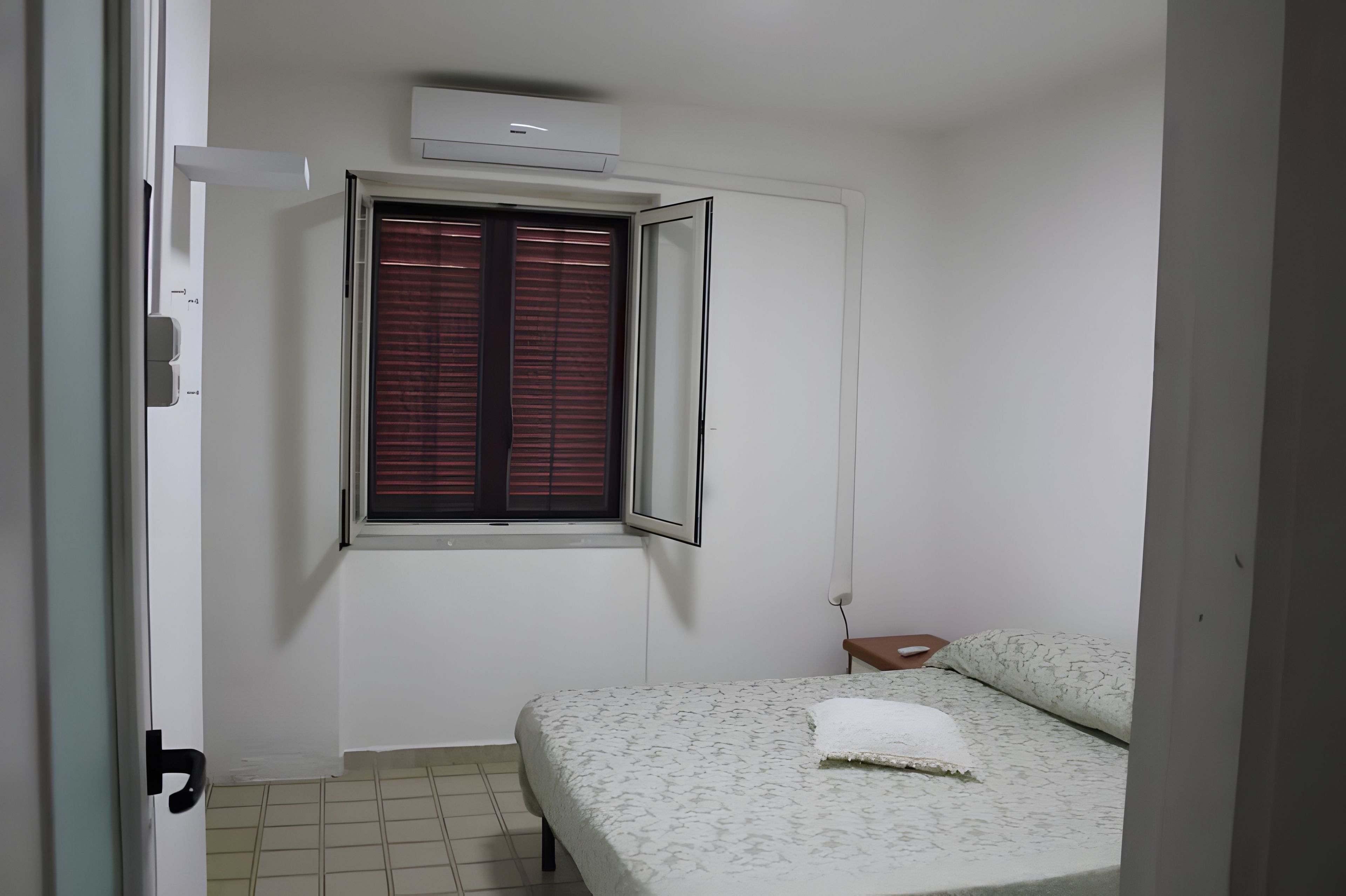 A small, neatly arranged bedroom with a bed, a window with blinds, a bedside table, and an air conditioning unit on the wall.