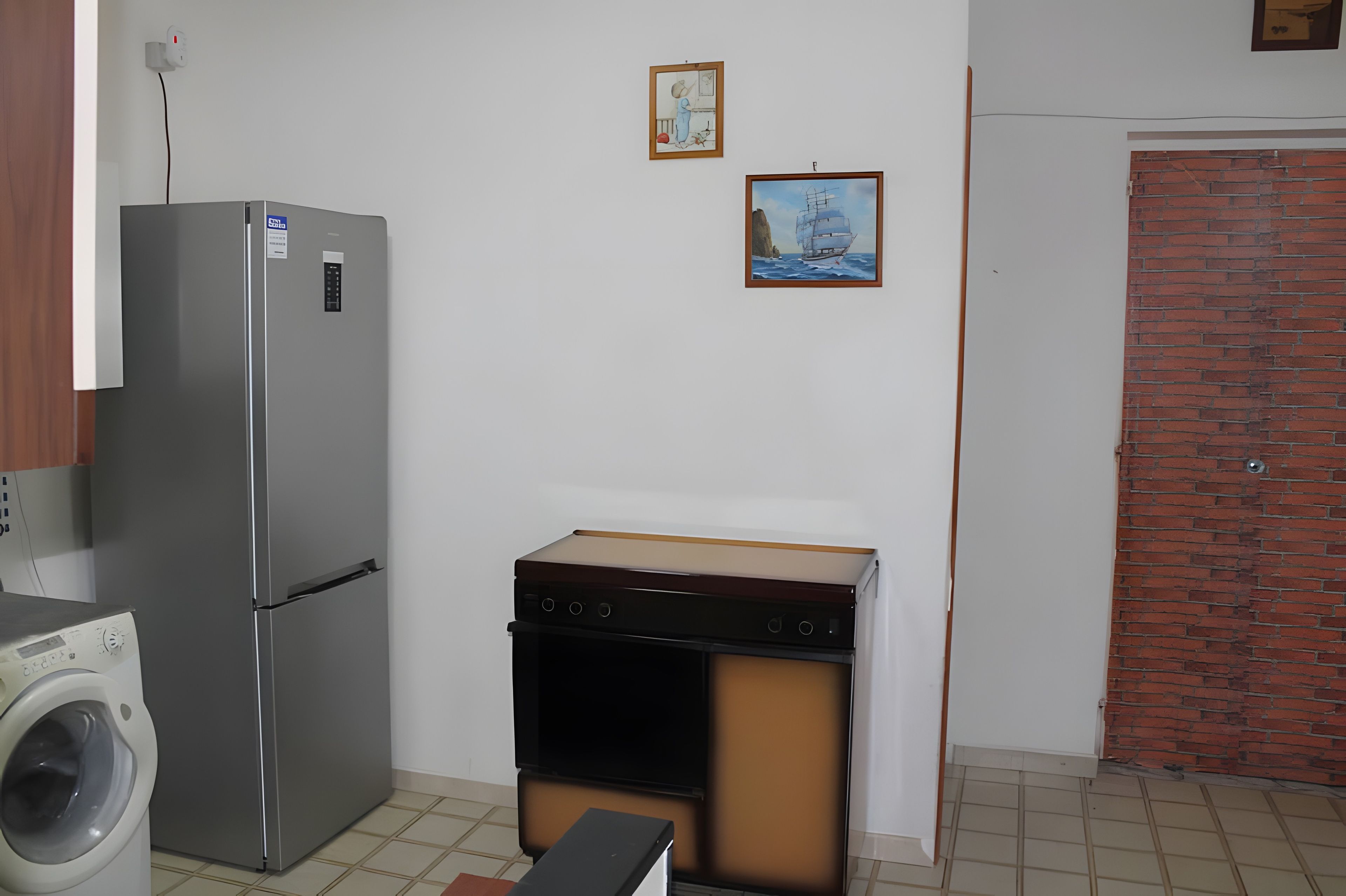 A compact kitchen with a refrigerator, washing machine, and a stove. There are two framed pictures on the wall.