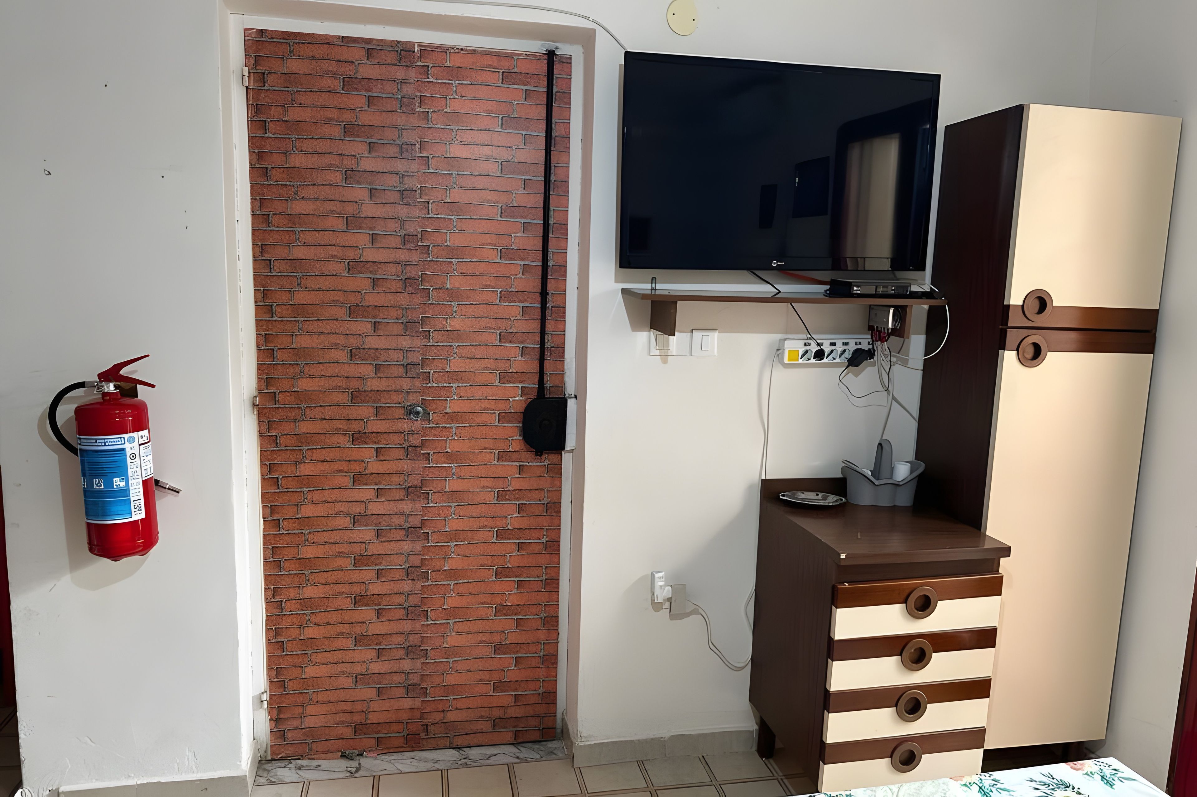 A room with a brick wall, a television mounted above a wooden cabinet, and a fire extinguisher on the wall.