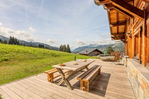 A wooden deck with a picnic table and benches, overlooking a grassy hillside with trees and a mountain in the distance. - Alpavi Chalet - Les Gets - Ski-in/Ski-out (Les Gets)