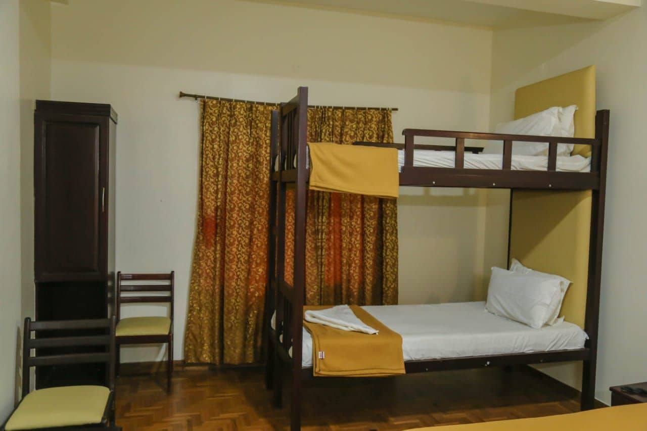 Una cama individual con dos literas de madera, una silla sencilla con cojín y una cortina con diseño estampado.