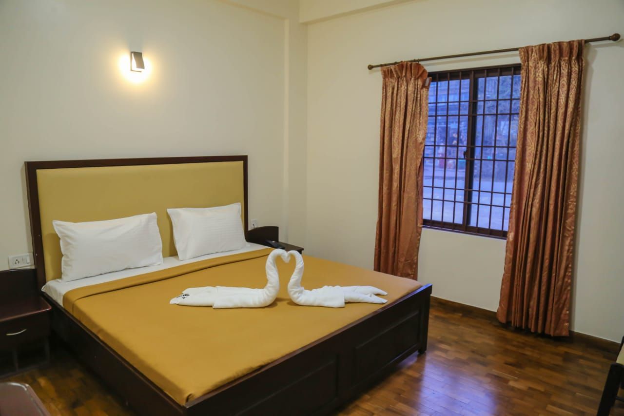 Una habitación de hotel con cama, mesita de noche, ventana con cortinas y una lámpara fijada a la pared.