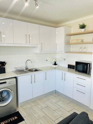 A modern kitchen with white cabinets, a built-in washing machine, a microwave, and a welcome mat. - Relax, Unwind & Feel at Home (Cape Town)