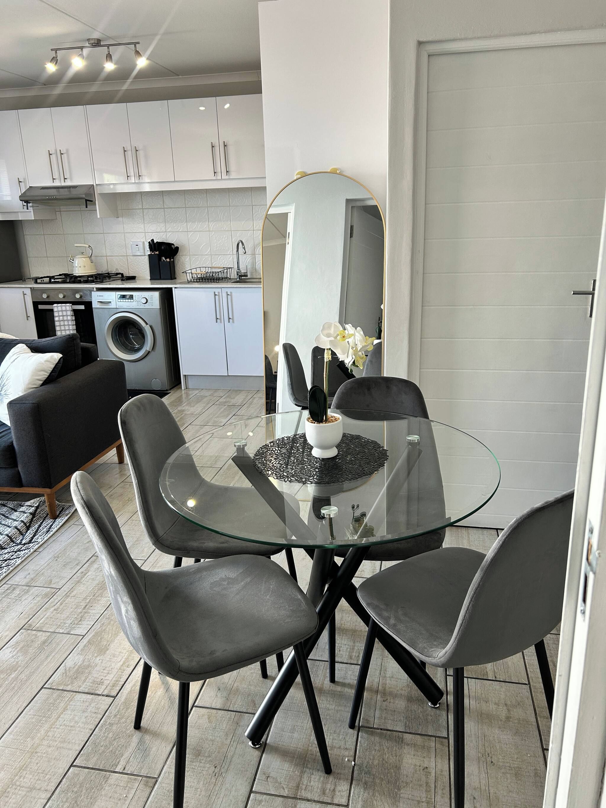 Een moderne keuken met witte kastjes, een wasmachine en een eethoek met een glazen tafel en stoelen.