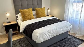 A modern bedroom with a grey upholstered headboard, a large bed with white and black bedding, two bedside lamps, and a grey rug.