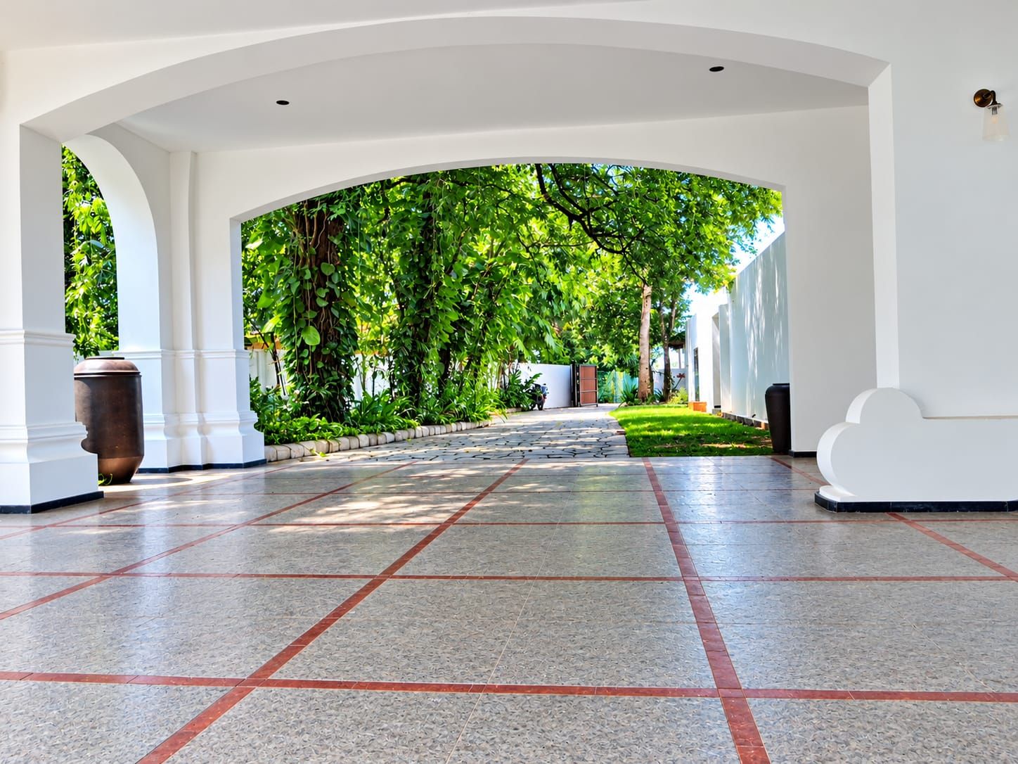 Un pasillo amplio con suelo de baldosas, columnas blancas y vistas a un jardín exuberante.