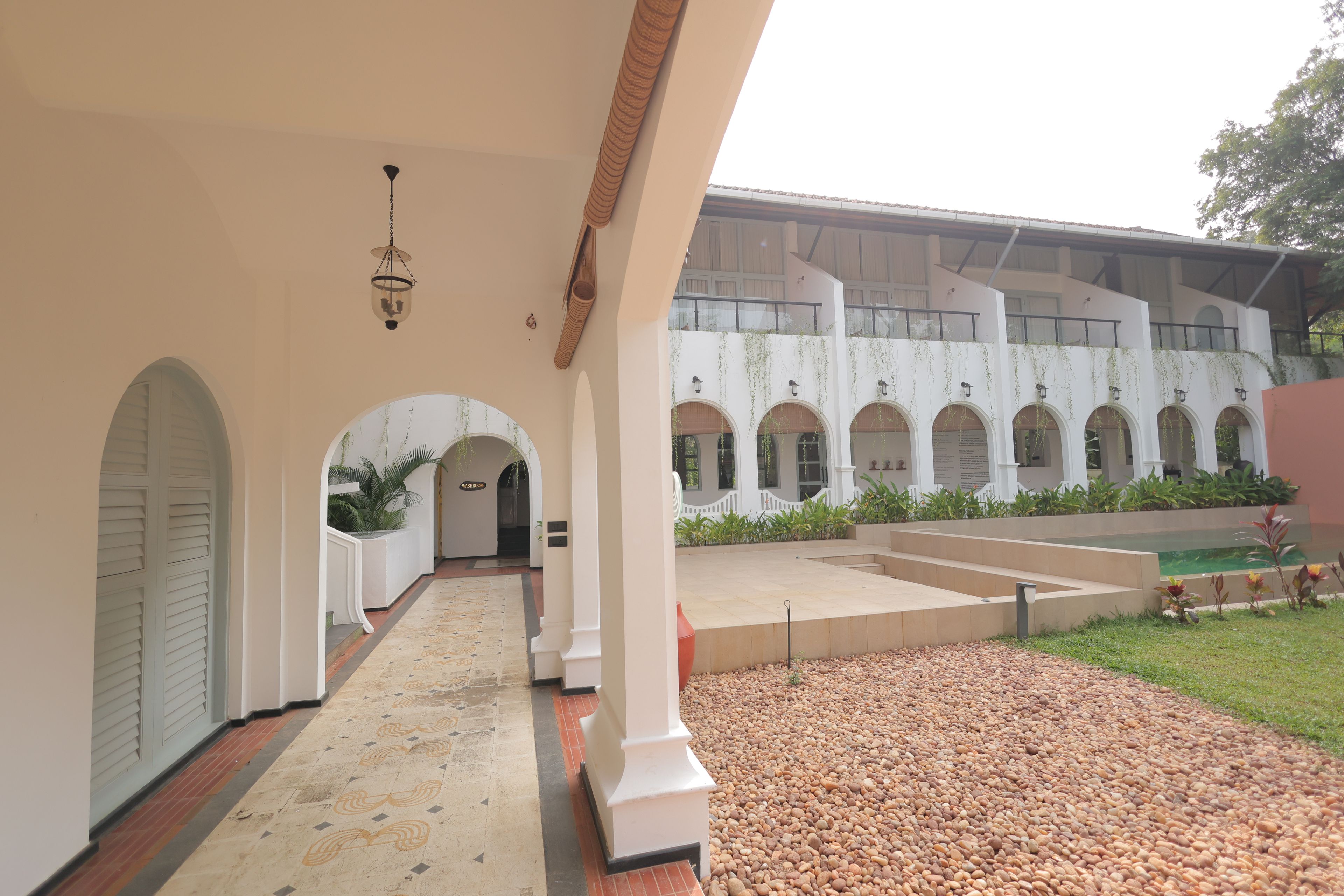 A covered walkway with arches, a tiled floor, and a building with multiple windows and a balcony.