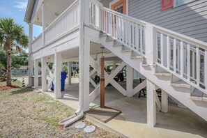 A covered outdoor area with a white staircase leading to a second level, surrounded by a concrete deck and a palm tree. - Barefoot Bungalow: Private Pool, Golf Cart Rental (Oak Island)