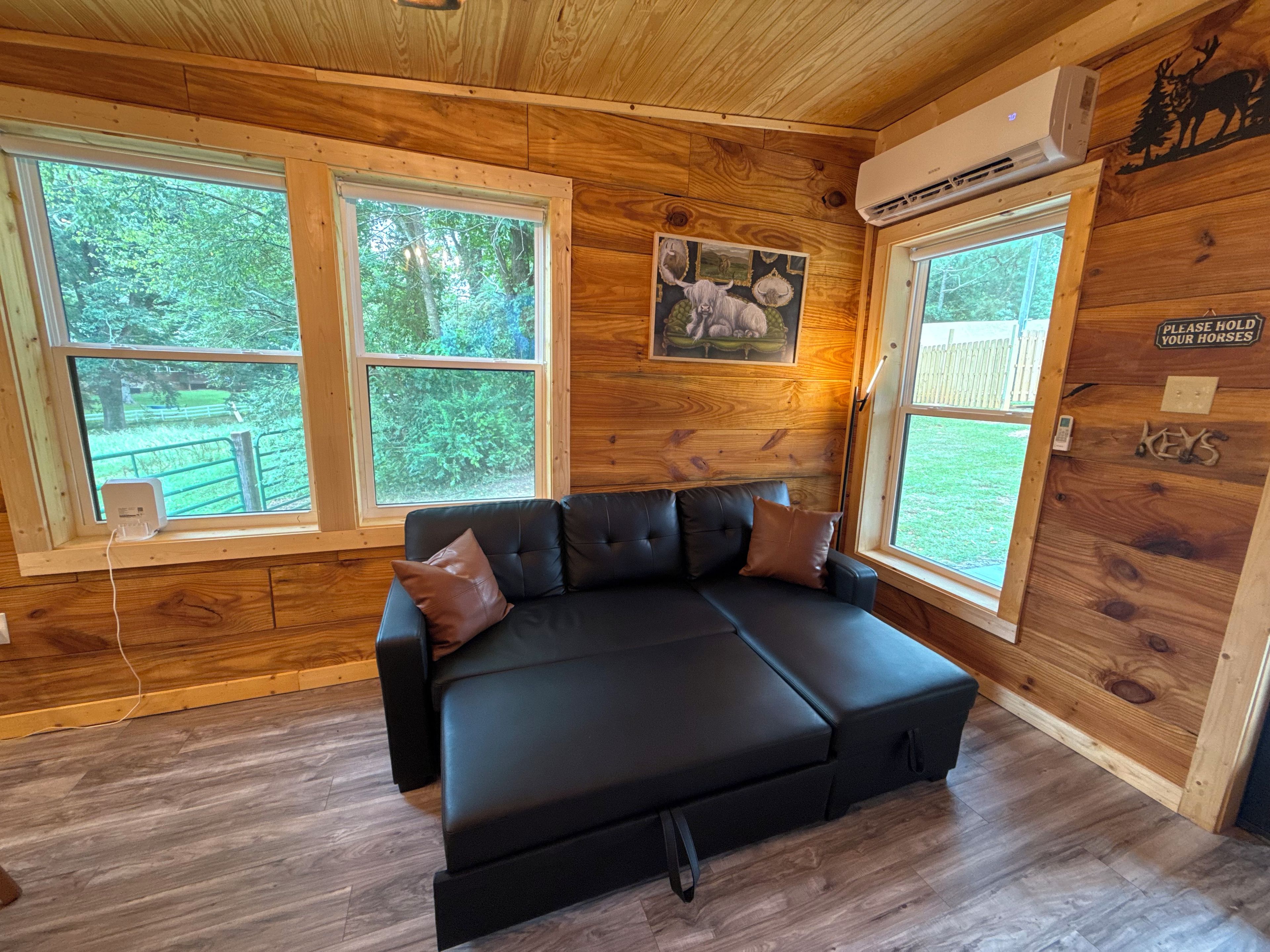 Interior de una cabaña de madera con un sofá de cuero negro, dos ventanas y un aire acondicionado de pared.