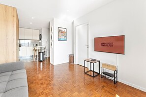 A modern hotel room with a wooden floor, a grey sofa, a small table with a laptop, a TV mounted on the wall, and a kitchen area in the background. - GuestReady - Cozy Porto Retreat with Balcony (Porto)