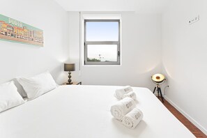 A hotel room with a bed, a nightstand with a lamp, a window with a view, and a wall with a cityscape painting. - GuestReady - Cozy Porto Retreat with Balcony (Porto)