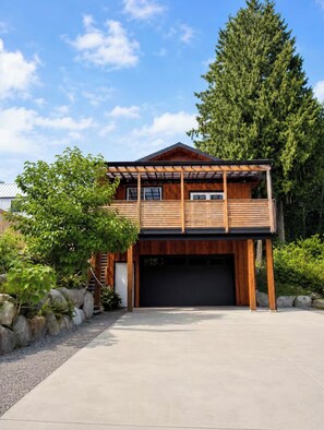 A two-story wooden house with a garage, surrounded by greenery and a clear sky. - Modern 1BR Suite in Granthams Landing (Gibsons)