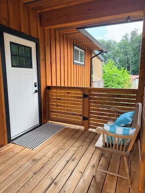 A wooden deck with a white door, a bench with a cushion, and a small window. - Modern 1BR Suite in Granthams Landing (Gibsons)