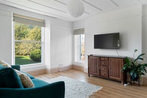 En stue med et stort vindue, et fladskærms-tv ophængt på væggen, et træskab og en teal-farvet sofa.
